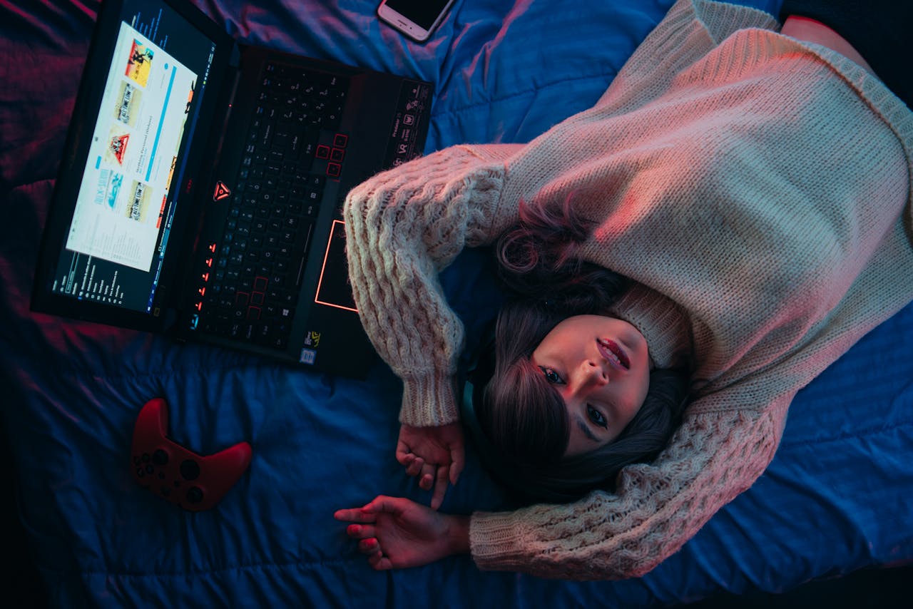 Teen girl lying on bed with laptop and game controller, perfect for gaming lifestyle visuals.